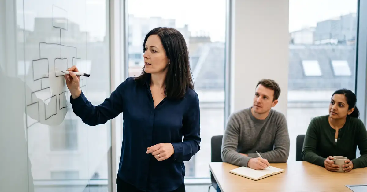 A UK head of sales sketching a team structure on a glass whiteboard while two colleagues listen
